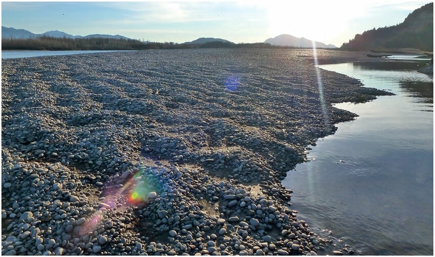 A large riverbed covered with mounds of smooth stones sits partially dry with water flowing along the edges, surrounded by mountains in the background.
